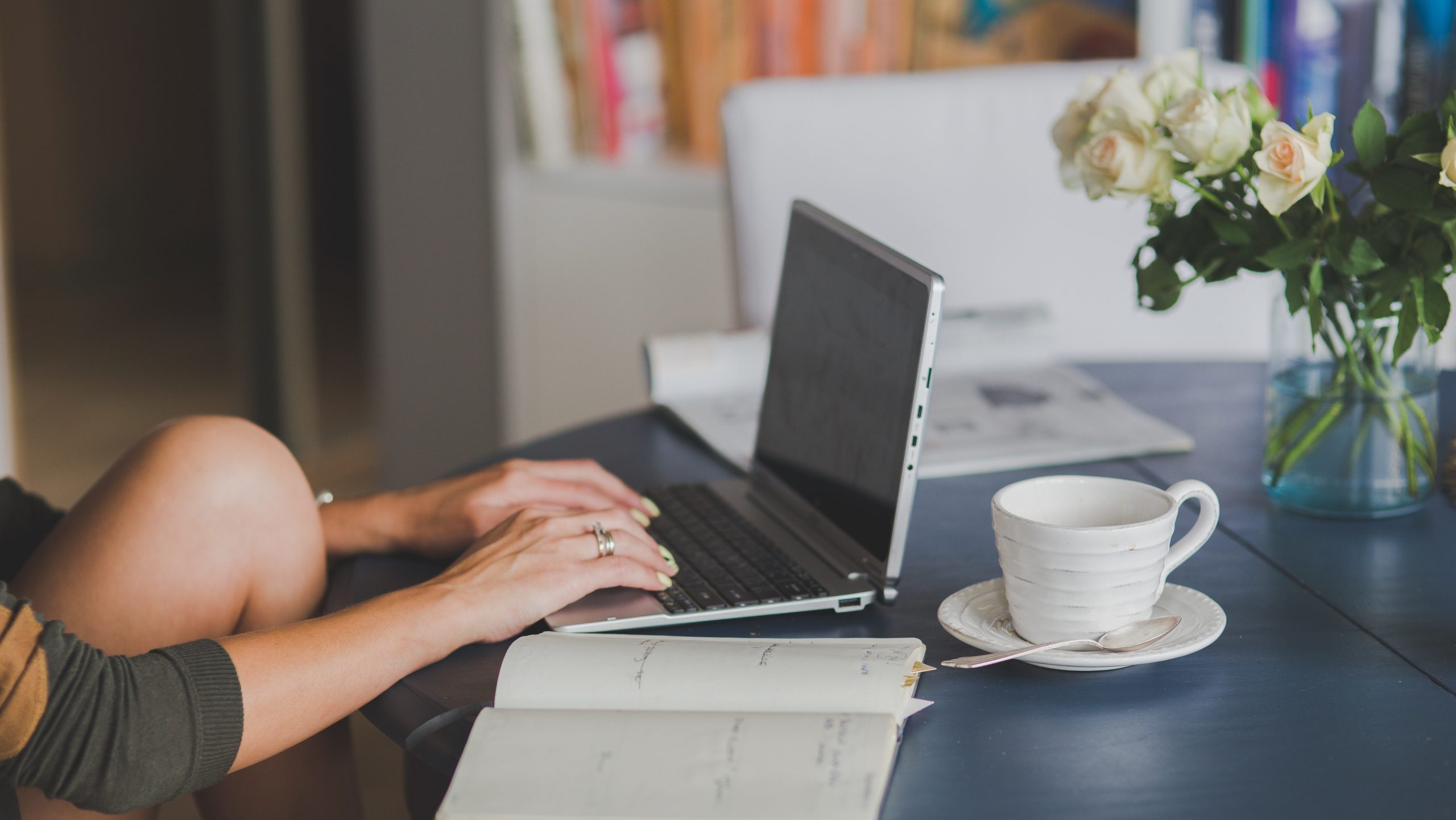 Image of the knee, arms, and hands of a woman sitting at a table working on a laptop, making her new website. These is a vase of roses and a coffee mug with saucer and spoon on the table.